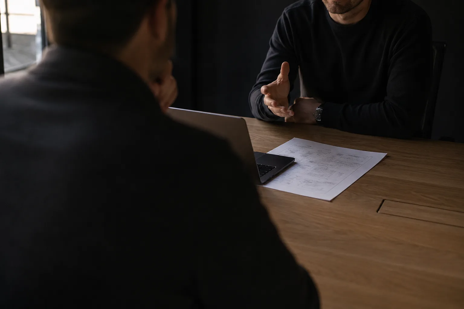 Seen over the shoulder of someone seated at a meeting table, facing another person explaining a point; a laptop and an annotated document lie between them.